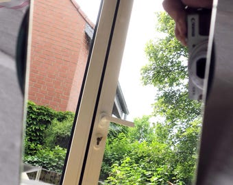 May include: A square mirror reflecting a view of a brick building, a white door, and lush green foliage. The mirror is held in front of a black object, with a camera visible in the reflection.