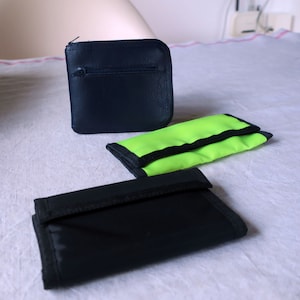 Könnte beinhalten: Drei kleine Geldbörsen, eine schwarze mit Reißverschluss, eine schwarze und eine leuchtend grüne.