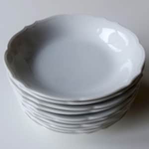 May include: A stack of white ceramic plates with a scalloped edge. The plates are stacked neatly on top of each other, with the top plate reflecting light. The plates have a simple, elegant design.