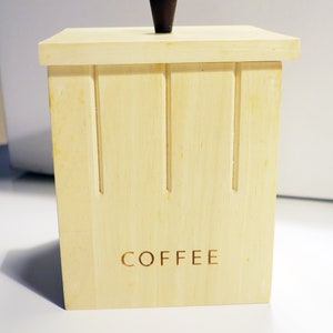 Peut inclure: Une boîte à café en bois avec un bouton brun foncé sur le couvercle. La boîte est de couleur brun clair et porte le mot "COFFEE" gravé sur le devant.