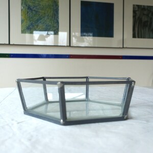 Könnte beinhalten: Ein sechseckiges Glas- und Metalltablett mit einem grauen Metallrahmen. Das Tablett ist leer und steht auf einer weißen Oberfläche.