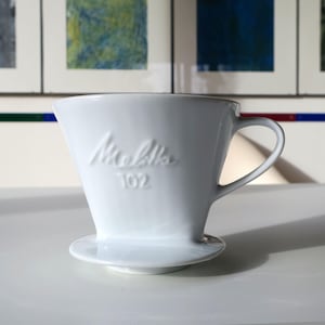 Peut inclure: Un filtre à café Melitta 102 en céramique blanche, avec une anse et une petite base. Le logo Melitta et "102" sont en relief sur le devant. Le filtre est conçu pour être placé sur une tasse ou une carafe pour infuser le café.