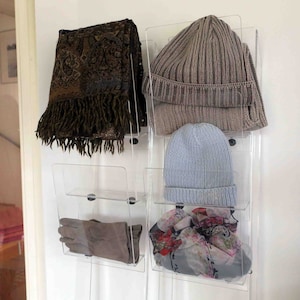 Könnte beinhalten: Ein transparenter, wandmontierter Organizer aus Acryl mit Regalen zur Aufbewahrung von Hüten, Schals und Handschuhen. Der Organizer hat vier Regale und ist mit einer Vielzahl von Winteraccessoires gefüllt, darunter ein brauner und goldener Schal, eine hellblaue Mütze, eine graue Mütze und ein floraler Schal.