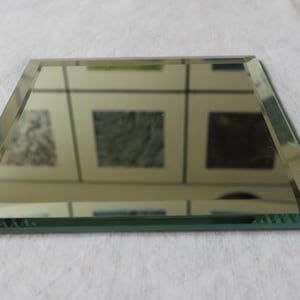 Può includere: Un piccolo specchio a forma di trapezio con bordi smussati. Lo specchio riflette l'ambiente circostante, comprese le opere d'arte incorniciate. Il vetro ha una leggera tinta verde e i bordi sono lucidi.