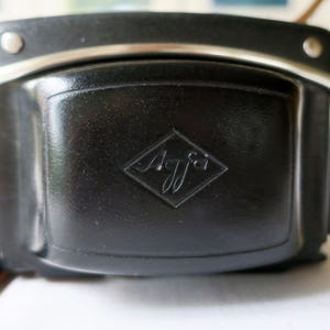 May include: A black leather camera case with a silver metal trim and strap attachments. The case features the embossed "Agfa" logo in a diamond shape on the front. The case is rectangular with rounded edges.