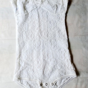 May include: White lace bodysuit with a halter neck. The one-piece garment features a floral lace pattern and a snap closure at the bottom. The edges are trimmed with a delicate, scalloped lace.
