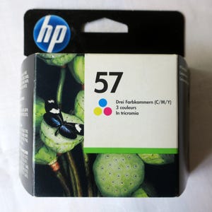 Könnte beinhalten: HP 57 Tintenpatrone in einer schwarz-weißen Verpackung. Die Verpackung zeigt einen Schmetterling auf einem grünen Lotus-Pod-Design. Der Text auf der Verpackung enthält "57", "Drei Farbkammern (C/M/Y)", "3 couleurs" und "In tricromia".