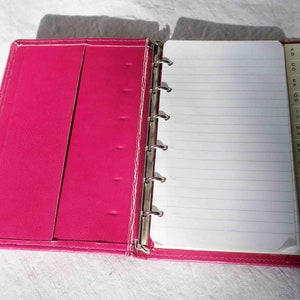 May include: A pink leather-bound notebook with a white lined paper insert and a tabbed index for alphabetical organisation.