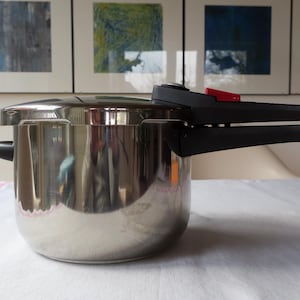 May include: A stainless steel pressure cooker with a black handle and a red release valve. The pot is sitting on a white surface, with artwork in the background.