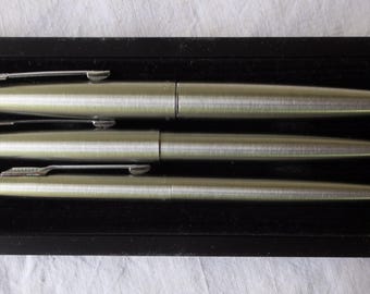 May include: A set of three silver-toned pens in a black presentation box. The pens have a sleek, metallic finish and are arranged neatly within the box. The pens are of varying sizes, suggesting a set of a pen, a pencil, and a smaller pen.