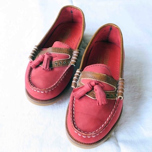 Könnte beinhalten: Ein Paar rote Lederschuhe mit braunen Ledereinsätzen und Quasten. Die Schuhe haben weiße Nähte an den Rändern.