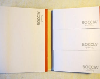 May include: A white notebook with a red and yellow spine. The front cover has the text "BOCCIA® TITANIUM" printed on it. The back cover has the text "BOCCIA® TITANIUM" printed on it four times.