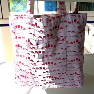 Può includere: Borsa tote bianca con motivo a villaggio rosso. La borsa presenta una tasca frontale con bottone e due manici. Il design include case e alberi su un motivo a linee ondulate.