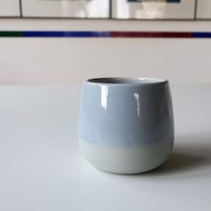 May include: A small, rounded ceramic cup with a two-tone design. The upper portion is a soft blue-gray, while the bottom half is a creamy white. The cup sits on a white surface.