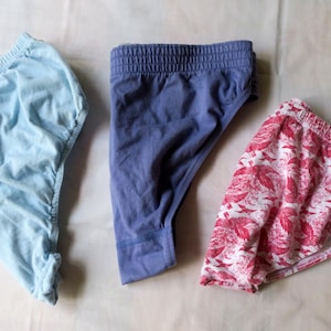 Op de afbeelding: Drie onderbroeken in verschillende kleuren en patronen. Eén is lichtblauw, een andere is effen donkerblauw en de derde heeft een witte achtergrond met een rood bloemenpatroon. Allemaal hebben ze elastische taillebanden.