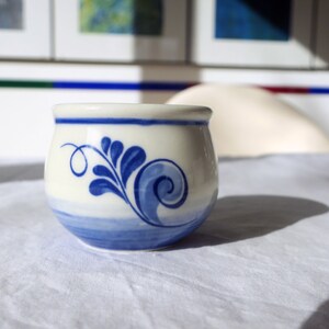 Puede incluir: Una taza de cerámica blanca con un diseño floral azul y blanco. La taza está sobre un mantel blanco.