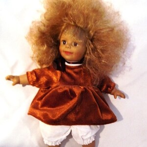 Könnte beinhalten: Eine Porzellanpuppe mit einem braunen Samtkleid und weißen Blümchen. Die Puppe hat blonde lockige Haare und braune Augen.