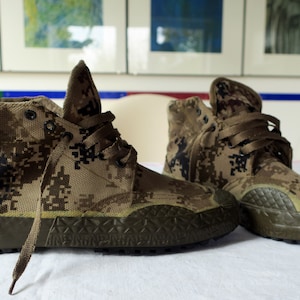 Könnte beinhalten: Ein Paar camouflagefarbene High-Top-Sneaker mit braunen Schnürsenkeln. Die Sneaker haben eine Gummisohle mit einem Profilmuster.