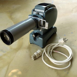 May include: Vintage gray slide projector with a long, cylindrical lens and a black lens cap. The projector has a rounded body and a base, with a power cord and plug. The projector is a classic piece of photography equipment.