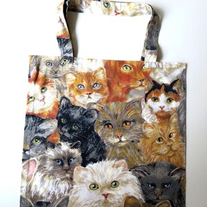 Può includere: Una borsa tote con un motivo ripetuto di facce di gatti nei toni dell'arancione, grigio, nero e bianco. I gatti hanno diversi colori degli occhi, tra cui blu e verde. La borsa ha uno sfondo bianco e manici abbinati.