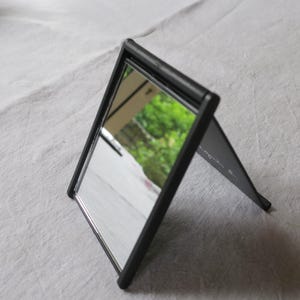 Puede incluir: Un espejo plegable rectangular negro. El espejo está abierto, reflejando una escena borrosa al aire libre. El marco es negro y el espejo es plateado. El espejo está sobre una superficie blanca.