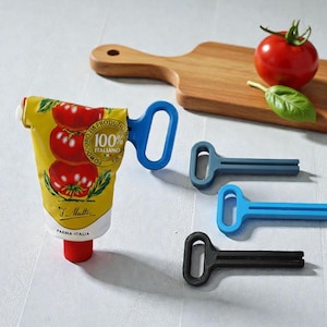 Könnte beinhalten: Eine gelbe Tomatenmarktube mit einem blauen, griffartigen Quetscher, zusammen mit drei weiteren Quetschern in Blau, Grau und Schwarz. Ein Holzschneidebrett, eine Tomate und ein Basilikumblatt sind ebenfalls vorhanden.