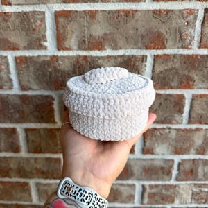 May include: A round, off-white crochet container with a matching lid, featuring a soft, textured yarn construction. The small, cylindrical vessel has a flat top and vertical sides, showcasing a consistent, dense stitch pattern throughout the design.