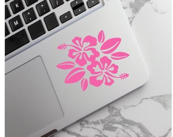 May include: A bright pink vinyl sticker featuring a graphic illustration of two hibiscus flowers with surrounding leaves. The sticker is applied to the silver surface of a laptop, positioned near the black keyboard keys and the trackpad area.