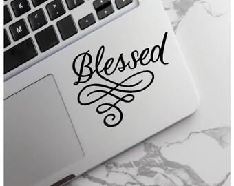 May include: A black vinyl decal sticker applied to the palm rest area of a silver laptop. The sticker features the word Blessed written in a black cursive script font, accompanied by an intricate, decorative flourish design underneath the text.
