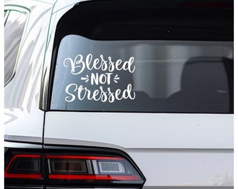 May include: A white vinyl decal applied to the rear window of a white vehicle. The decal features the text "Blessed NOT Stressed" in a stylized, cursive script font, with small decorative flourishes flanking the word "NOT" in the center.