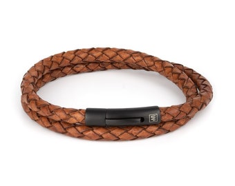 Puede incluir: Un brazalete de cuero marrón con un diseño trenzado y un cierre de metal negro. El cierre tiene un pequeño logotipo de metal con las letras "WM".