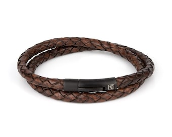 Puede incluir: Un brazalete de cuero marrón con un diseño trenzado y un cierre de metal negro. El cierre tiene una pequeña placa de metal con la letra "W" en ella.