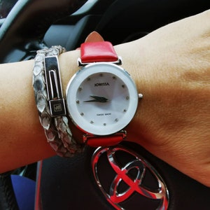 May include: A silver watch with a white face and red leather strap. The watch face has a silver JOWISSA logo and the words "SWISS MADE". The watch is on a person's wrist with a silver snake skin bracelet.