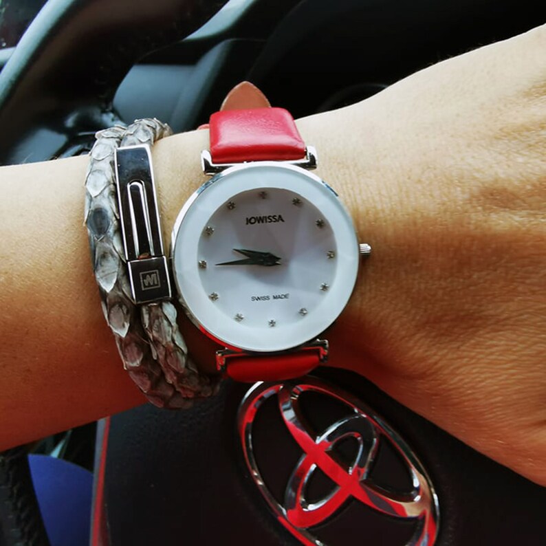 May include: A silver watch with a white face and red leather strap. The watch face has a silver JOWISSA logo and the words "SWISS MADE". The watch is on a person's wrist with a silver snake skin bracelet.