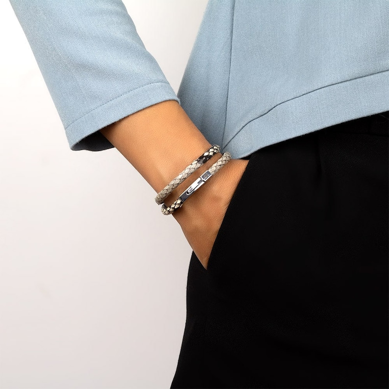 May include: Two braided leather bracelets, one black and one light grey, with silver clasps. The bracelets are worn on a person's wrist.