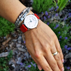 May include: A silver watch with a white face and diamond accents. The watch has a red leather strap and is worn on a wrist with a silver bracelet.