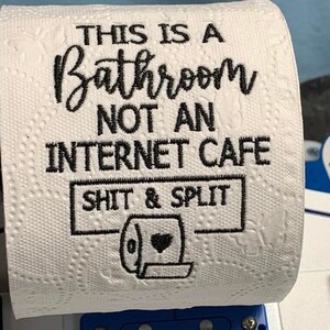 K&ouml;nnte beinhalten: Wei&szlig;e Toilettenpapierrolle mit schwarzem Text: &bdquo;THIS IS A BATHROOM NOT AN INTERNET CAFE SHIT & SPLIT&ldquo; und einer Toilettengrafik. Die Rolle ist auf einem blauen Kunststoffhalter montiert.