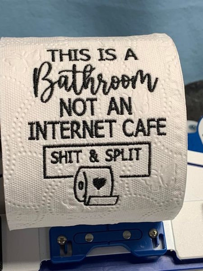 K&ouml;nnte beinhalten: Wei&szlig;e Toilettenpapierrolle mit schwarzem Text: &bdquo;THIS IS A BATHROOM NOT AN INTERNET CAFE SHIT & SPLIT&ldquo; und einer Toilettengrafik. Die Rolle ist auf einem blauen Kunststoffhalter montiert.