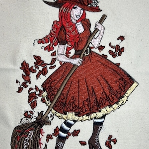 May include: Embroidered design of a witch with red hair wearing a brown witch hat and a red dress, holding a broom and sweeping up brown leaves.