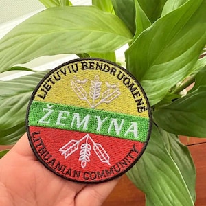 May include: A round embroidered patch featuring the Lithuanian flag colors: yellow, green, and red. The patch includes the text "LIETUVIŲ BENDRUOMENĖ" and "ŽEMYNA" in white, along with a wheat stalk design. "LITHUANIAN COMMUNITY" is also displayed.
