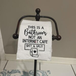 K&ouml;nnte beinhalten: Wei&szlig;e Toilettenpapierrolle mit dem Text "THIS IS A BATHROOM NOT AN INTERNET CAFE SHIT & SPLIT" in schwarzer Schrift. Die Rolle befindet sich auf einem dunkelbraunen Toilettenpapierhalter aus Metall.