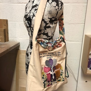 May include: A beige tote bag with a floral design and the text "The best things in life are not things, they are sisters". The bag has a long strap and a drawstring closure.