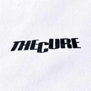 May include: White fabric with the embroidered text "THE CURE" in black, angled diagonally across the image. The text has a slightly distressed, textured appearance, suggesting an embroidered design.