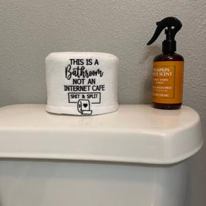 K&ouml;nnte beinhalten: Eine wei&szlig;e Toilettenpapierabdeckung mit dem Text "THIS IS A Bathroom NOT AN INTERNET CAFE SHIT & SPLIT" und einer Toilettenpapierrolle. Daneben eine braune Flasche Raumspray "PUMPKIN SPICE SCENT".