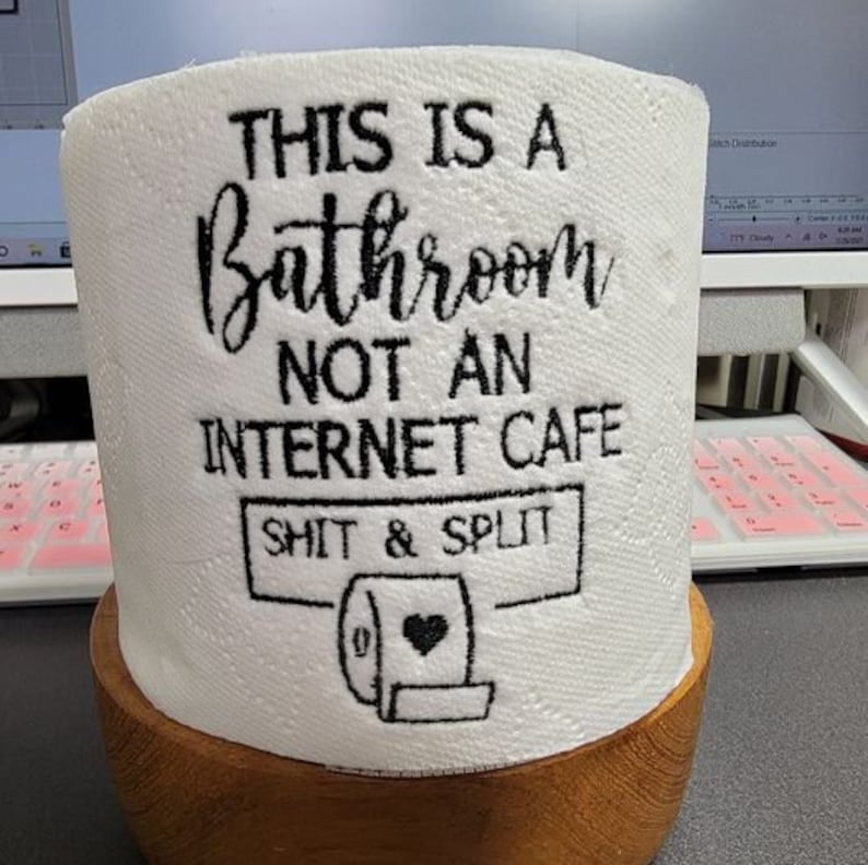 K&ouml;nnte beinhalten: Wei&szlig;e Toilettenpapierrolle mit schwarzem, gesticktem Text: "THIS IS A BATHROOM NOT AN INTERNET CAFE SHIT & SPLIT" und einer Grafik einer Toilettenpapierrolle mit Herz. Die Rolle steht in einem Holzhalter.