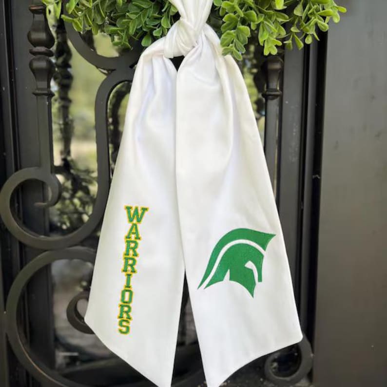 May include: White sash with the word "Warriors" in green and gold lettering. A green Spartan helmet logo is on the other side. The sash is tied at the top and hangs down. The sash is hanging on a black wrought iron gate.