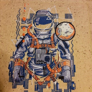 May include: Embroidered astronaut design in shades of blue, orange, and gray. The astronaut is wearing a spacesuit with a detailed helmet and harness. A planet with craters is depicted to the right. The background is a tan fabric with a quilted pattern.