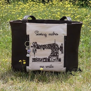 Puede incluir: Un bolso tote marr&oacute;n con asas negras y un panel frontal color crema. El panel presenta una ilustraci&oacute;n negra de una m&aacute;quina de coser y el texto "Sewing makes me smile". Un llavero con abalorios cuelga del lateral.