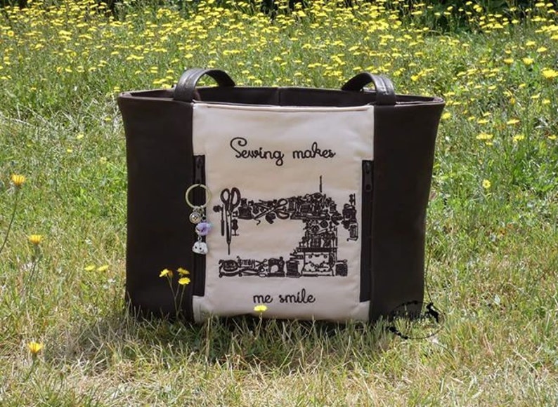 Puede incluir: Un bolso tote marr&oacute;n con asas negras y un panel frontal color crema. El panel presenta una ilustraci&oacute;n negra de una m&aacute;quina de coser y el texto "Sewing makes me smile". Un llavero con abalorios cuelga del lateral.