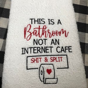 K&ouml;nnte beinhalten: Wei&szlig;es Badezimmer-Handtuch mit gesticktem Text: "THIS IS A Bathroom NOT AN INTERNET CAFE SHIT & SPLIT" und einer Toilettenpapierrolle mit rotem Herz. Das Handtuch liegt auf einer schwarz-wei&szlig; karierten Oberfl&auml;che.